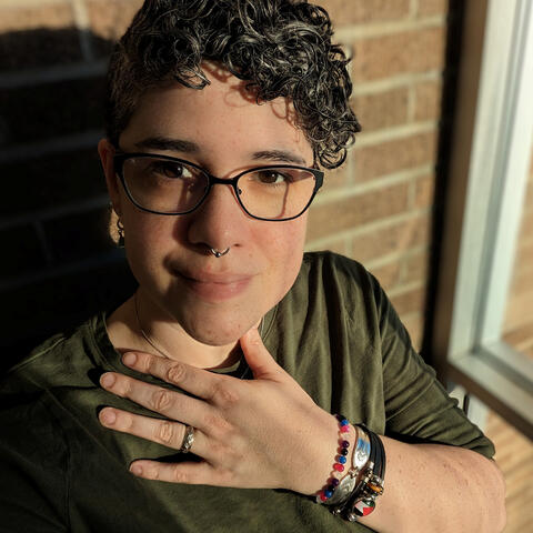 Nonbinary individual standing against a brick wall, their hand resting on their chest.